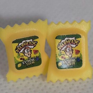 May include: A pair of yellow Warheads candy earrings. The earrings are shaped like a small candy packet with the Warheads logo and the words "Extreme Sour".