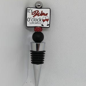 May include: A silver and black wine bottle stopper with a red and white sign that reads "It's Wine O'clock Somewhere".