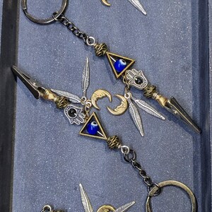 May include: Keychains with a silver spearhead design, adorned with a blue evil eye in a triangle, a gold crescent moon, and a Hamsa hand charm. The keychains have a silver chain and a gold-colored key ring.