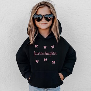 May include: A black hooded sweatshirt with the text "favorite daughter" in pink script, surrounded by pink bow graphics. The sweatshirt has a front pocket and is worn by a young person wearing heart-shaped sunglasses.
