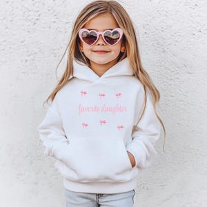 May include: A white hoodie with a front pocket and the text "favorite daughter" in pink. The hoodie has pink bow graphics and is worn with pink heart-shaped sunglasses.