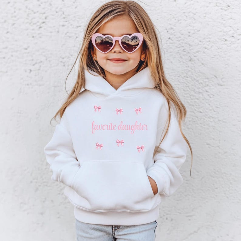May include: A white hoodie with a front pocket and the text "favorite daughter" in pink. The hoodie has pink bow graphics and is worn with pink heart-shaped sunglasses.