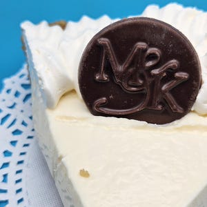 May include: A slice of pie with a creamy white filling and whipped cream topping. A dark chocolate disc with the letters "MK" is on top. The pie sits on a white doily against a blue background.
