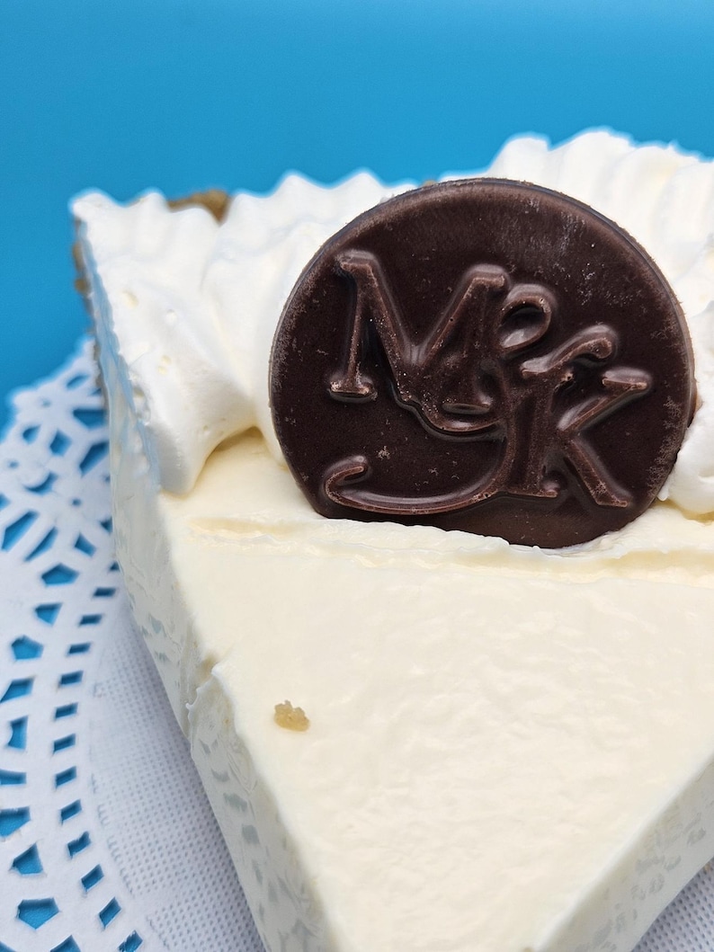 May include: A slice of pie with a creamy white filling and whipped cream topping. A dark chocolate disc with the letters "MK" is on top. The pie sits on a white doily against a blue background.