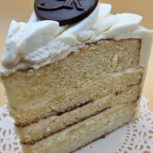 May include: A slice of layered cake with white frosting and a dark chocolate disc on top. The cake has three visible layers of light-colored sponge cake with a creamy filling. The frosting is swirled, and the chocolate disc has an embossed design.