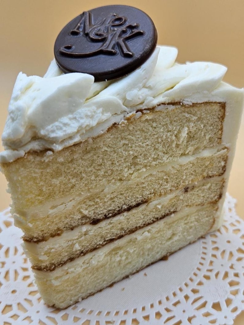 May include: A slice of layered cake with white frosting and a dark chocolate disc on top. The cake has three visible layers of light-colored sponge cake with a creamy filling. The frosting is swirled, and the chocolate disc has an embossed design.