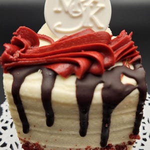 May include: A red velvet cupcake with white frosting, red frosting swirls, and chocolate drizzle. A white circular decoration with the letters "N&K" sits atop the cupcake. The cupcake sits on a white paper doily.
