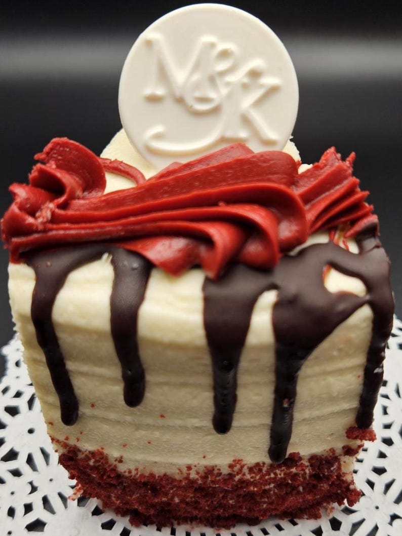 May include: A red velvet cupcake with white frosting, red frosting swirls, and chocolate drizzle. A white circular decoration with the letters "N&K" sits atop the cupcake. The cupcake sits on a white paper doily.