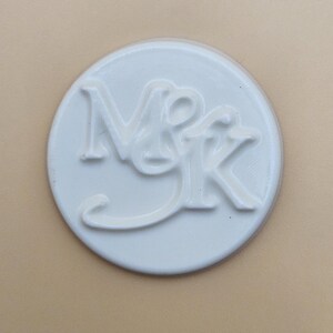 May include: A white, circular object with the embossed letters "NMK". The letters are in a stylized font, with the "M" and "K" connected. The object is set against a neutral, light tan background.