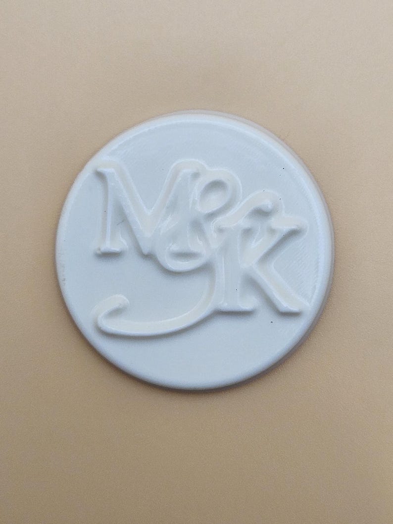 May include: A white, circular object with the embossed letters "NMK". The letters are in a stylized font, with the "M" and "K" connected. The object is set against a neutral, light tan background.