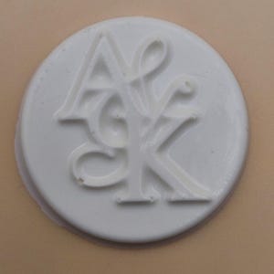 May include: A white, circular wax seal with the intertwined letters "A", "S", and "K" in a stylized font. The seal is set against a light tan background, creating a contrast that highlights the embossed design.