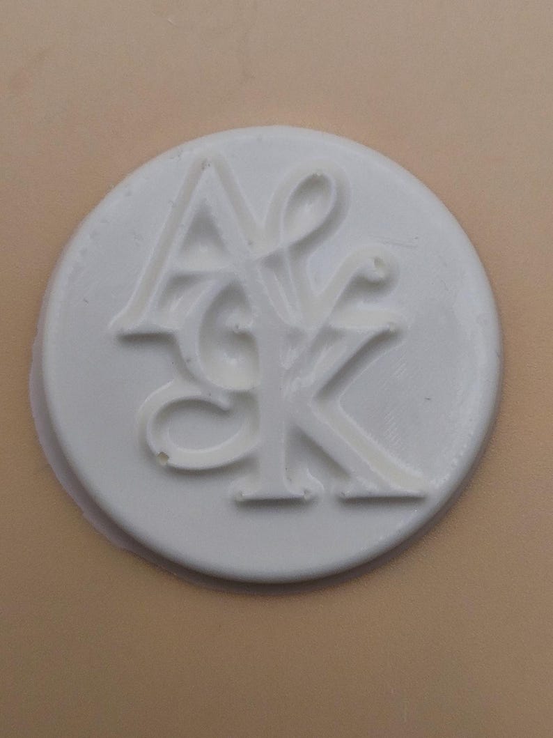 May include: A white, circular wax seal with the intertwined letters "A", "S", and "K" in a stylized font. The seal is set against a light tan background, creating a contrast that highlights the embossed design.