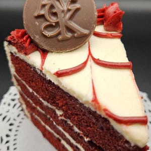 May include: A slice of red velvet cake with white frosting and red accents. A chocolate disc with the letters "AK" is on top. The cake sits on a white paper doily.