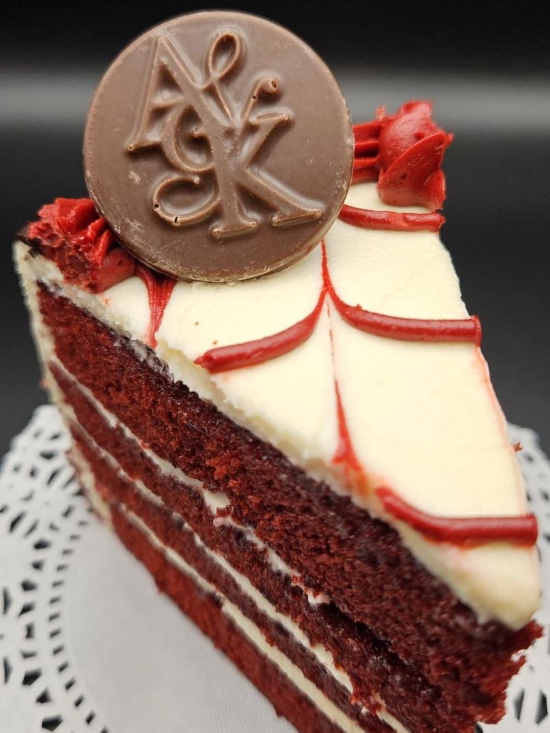 May include: A slice of red velvet cake with white frosting and red accents. A chocolate disc with the letters "AK" is on top. The cake sits on a white paper doily.