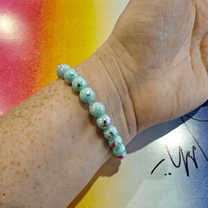 May include: A beaded bracelet with turquoise-colored beads and red accents is worn on a wrist. The bracelet is made of round beads. The background is a colorful abstract design with pink, yellow, and blue hues.
