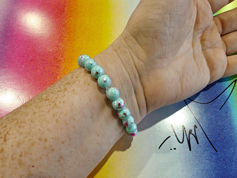 May include: A beaded bracelet with turquoise-colored beads and red accents is worn on a wrist. The bracelet is made of round beads. The background is a colorful abstract design with pink, yellow, and blue hues.