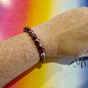 May include: A beaded bracelet with red and silver beads is worn on a freckled arm. The bracelet is made of round beads with a textured, patterned surface. The background is a colorful gradient of pink, yellow, and blue.
