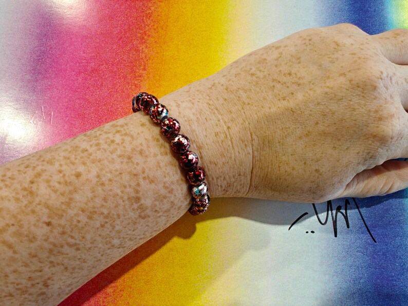 May include: A beaded bracelet with red and silver beads is worn on a freckled arm. The bracelet is made of round beads with a textured, patterned surface. The background is a colorful gradient of pink, yellow, and blue.