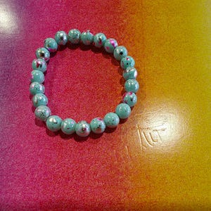 May include: A turquoise beaded bracelet with a speckled design. The bracelet is made of round beads and is displayed on a colorful background with shades of pink, red, and yellow.