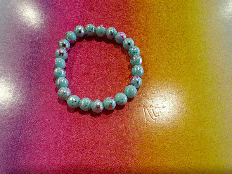 May include: A turquoise beaded bracelet with a speckled design. The bracelet is made of round beads and is displayed on a colorful background with shades of pink, red, and yellow.