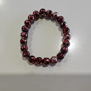 May include: A beaded bracelet featuring round beads with a speckled design in shades of red, white, and blue. The bracelet is made of elastic and is designed to be worn on the wrist. The beads are approximately 1 cm in diameter.