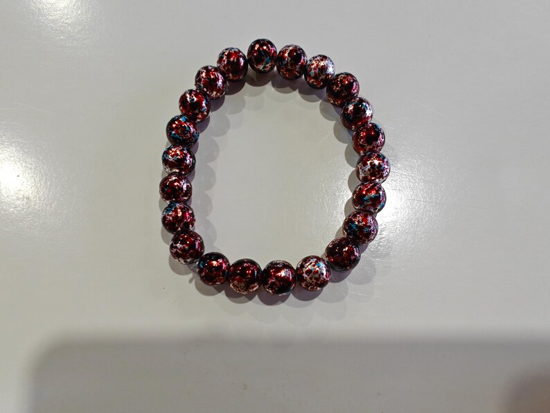 May include: A beaded bracelet featuring round beads with a speckled design in shades of red, white, and blue. The bracelet is made of elastic and is designed to be worn on the wrist. The beads are approximately 1 cm in diameter.