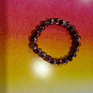 May include: A beaded bracelet with dark red and iridescent beads. The bracelet is circular and sits on a gradient background of yellow, orange, and pink.