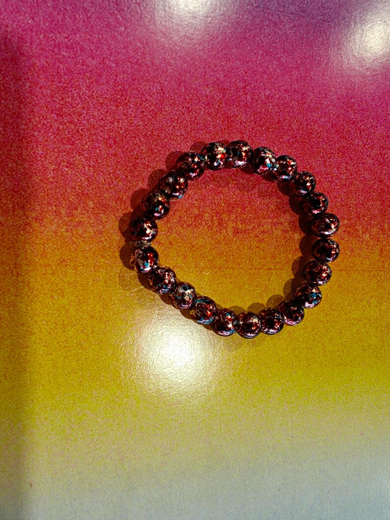 May include: A beaded bracelet with dark red and iridescent beads. The bracelet is circular and sits on a gradient background of yellow, orange, and pink.