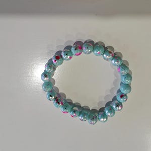 May include: A beaded bracelet featuring light blue, round beads with pink and red accents. The bracelet is made of elastic and is designed to be worn around the wrist. The beads have a glossy finish.