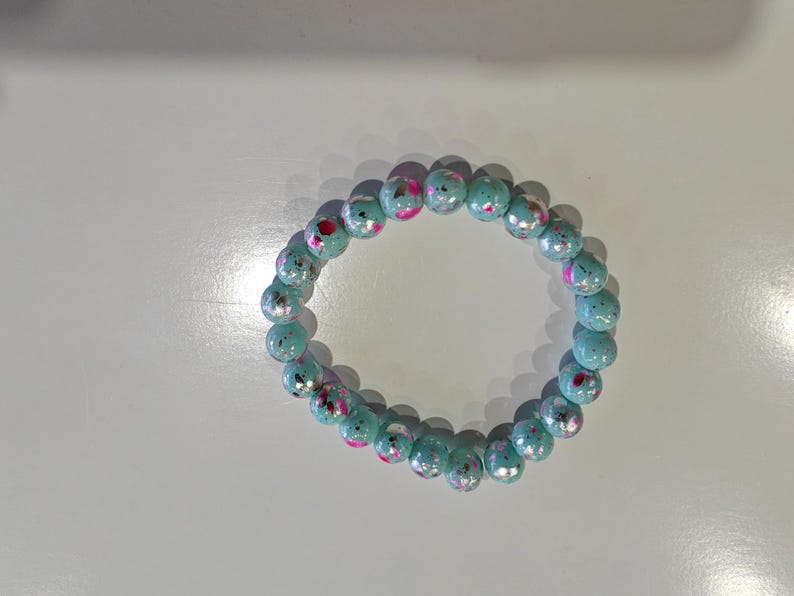 May include: A beaded bracelet featuring light blue, round beads with pink and red accents. The bracelet is made of elastic and is designed to be worn around the wrist. The beads have a glossy finish.