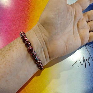 May include: A beaded bracelet with red, black, and blue accents. The bracelet is made of round beads and is worn on a wrist. The background is a colorful gradient of pink, yellow, and blue.