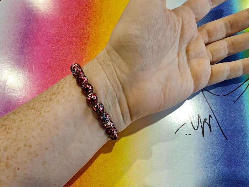 May include: A beaded bracelet with red, black, and blue accents. The bracelet is made of round beads and is worn on a wrist. The background is a colorful gradient of pink, yellow, and blue.