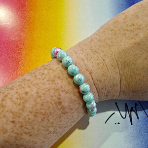 May include: A beaded bracelet with turquoise and pink speckled beads. The bracelet is worn on a freckled arm against a colorful background. The beads are approximately 0.5 inches in diameter. The bracelet is a piece of jewelry.