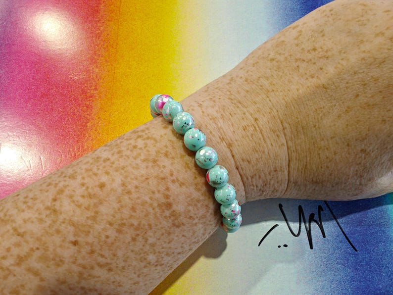 May include: A beaded bracelet with turquoise and pink speckled beads. The bracelet is worn on a freckled arm against a colorful background. The beads are approximately 0.5 inches in diameter. The bracelet is a piece of jewelry.