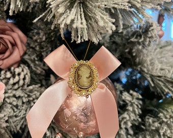May include: A pink glass Christmas ornament with a pink satin bow and a gold cameo brooch. The ornament is decorated with small, clear beads.