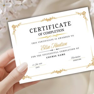 Op de afbeelding: Een wit certificaat met gouden bloemaccenten en zwarte tekst. Het certificaat luidt "CERTIFICATE OF COMPLETION" en is uitgereikt aan "Elsa Hudson". De datum is 23 januari 2036. Het certificaat wordt door een hand vastgehouden.