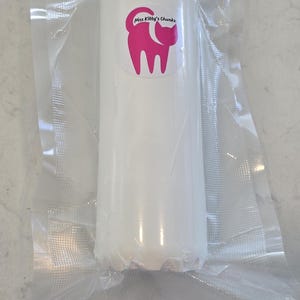 May include: A white, cylindrical product, vacuum-sealed in clear plastic. A pink sticker with the text "Miss Kitty's Chunks" and a cat silhouette is visible on the product. The product is on a light gray surface.