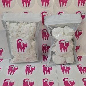 May include: Two clear plastic pouches filled with white edible chunks, positioned over a sheet of circular stickers featuring a pink cat silhouette and the text Miss Kitty's Chunkz. The left pouch contains small, irregular pieces, while the right pouch holds larger, rounded white pieces.