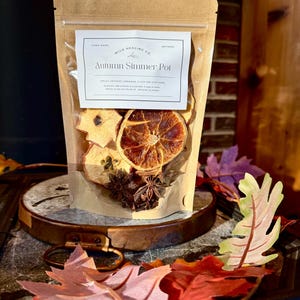 Könnte beinhalten: Ein klarer Beutel gefüllt mit getrockneten Apfel- und Orangenscheiben, Sternanis und Nelken. Das Etikett trägt die Aufschrift "Autumn Simmer Pot". Der Beutel liegt auf einer Holzoberfläche, umgeben von Herbstblättern.