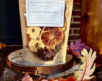May include: A clear bag filled with dried apple slices, orange slices, star anise, and cloves. The bag has a label that reads "Autumn Simmer Pot" and is sitting on a wooden surface with fall leaves.