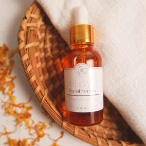 K&ouml;nnte beinhalten: Eine Glasflasche mit Gesichts-Serum mit goldenem Deckel und Tropfer. Die Flasche ist mit "GLOW Facial Serum" beschriftet und befindet sich in einem geflochtenen Korb. Die Flasche hat ein Fassungsverm&ouml;gen von 1 fl oz.