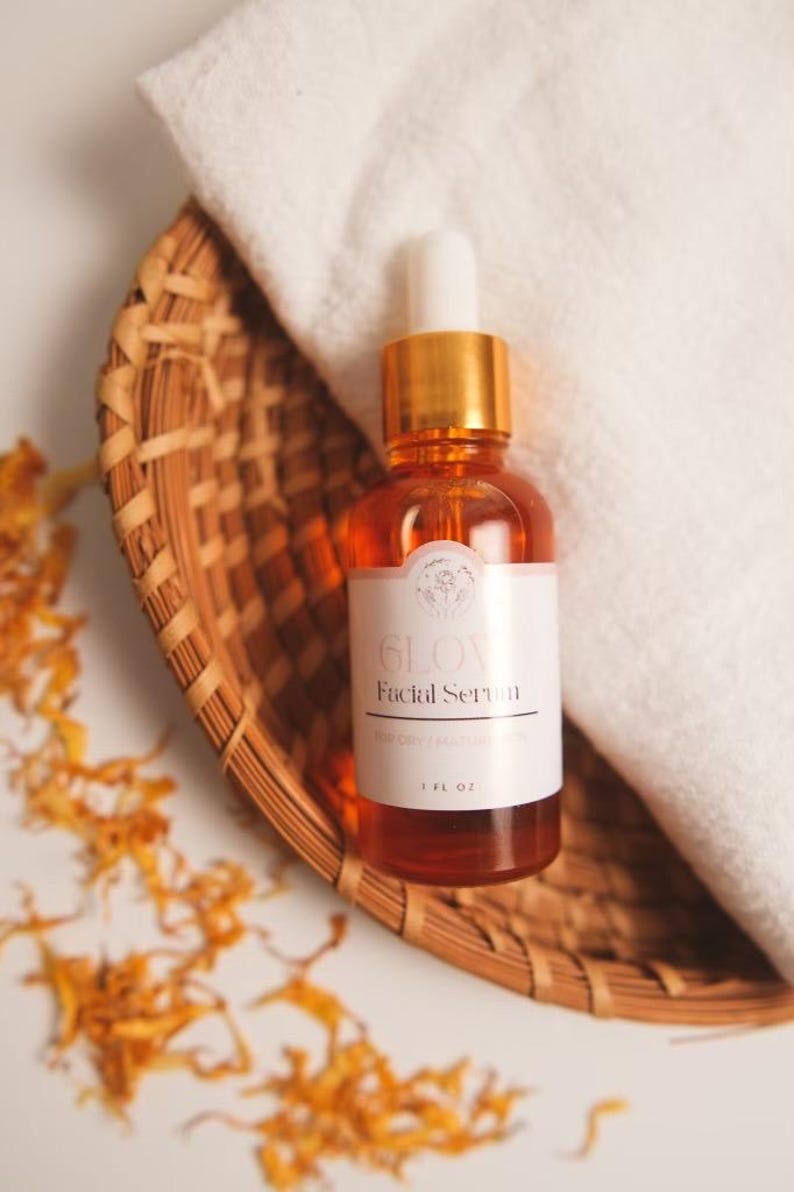 K&ouml;nnte beinhalten: Eine Glasflasche mit Gesichts-Serum mit goldenem Deckel und Tropfer. Die Flasche ist mit "GLOW Facial Serum" beschriftet und befindet sich in einem geflochtenen Korb. Die Flasche hat ein Fassungsverm&ouml;gen von 1 fl oz.