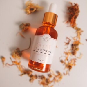 K&ouml;nnte beinhalten: Eine Glasflasche mit bernsteinfarbenem Gesichts-Serum mit goldenem Deckel und Tropfer. Die Flasche ist mit "GLOW Facial Serum" und "1 FL OZ" beschriftet. Die Flasche ist von getrockneten orangefarbenen Bl&uuml;tenbl&auml;ttern auf wei&szlig;em Hintergrund umgeben.