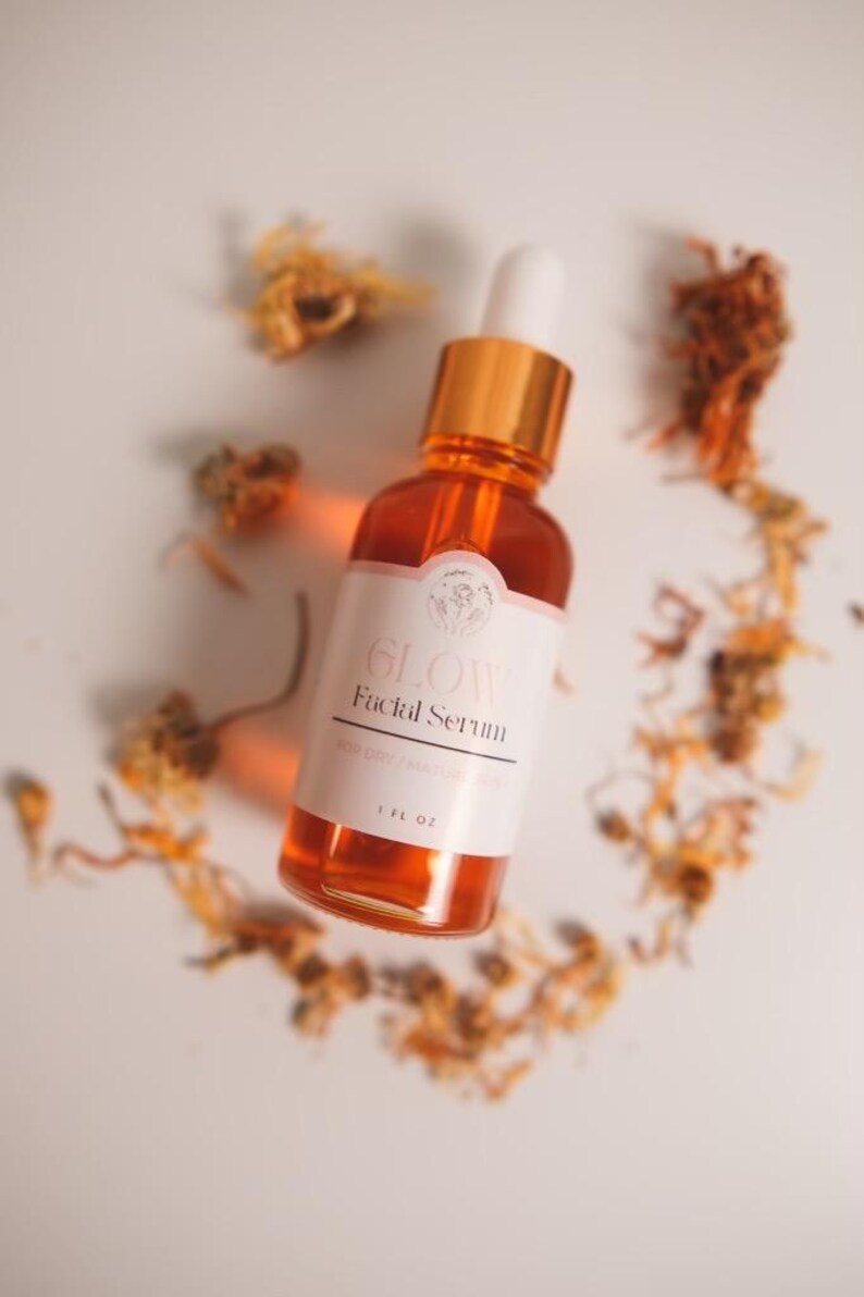 K&ouml;nnte beinhalten: Eine Glasflasche mit bernsteinfarbenem Gesichts-Serum mit goldenem Deckel und Tropfer. Die Flasche ist mit "GLOW Facial Serum" und "1 FL OZ" beschriftet. Die Flasche ist von getrockneten orangefarbenen Bl&uuml;tenbl&auml;ttern auf wei&szlig;em Hintergrund umgeben.