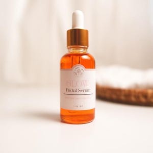 K&ouml;nnte beinhalten: Eine Glasflasche mit Gesichts-Serum mit goldenem Deckel und Tropfer. Die Flasche ist mit "Glow Facial Serum" beschriftet und enth&auml;lt eine klare orange Fl&uuml;ssigkeit. Die Flasche steht auf einer wei&szlig;en Oberfl&auml;che.