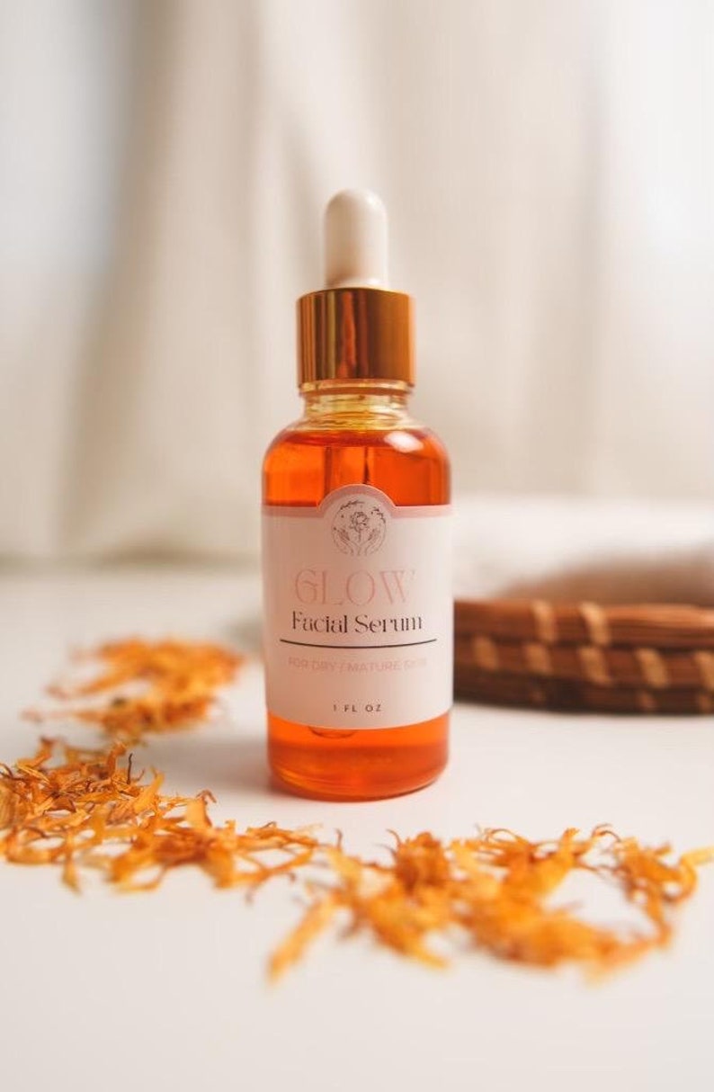K&ouml;nnte beinhalten: Eine Glasflasche mit Gesichts-Serum mit goldenem Deckel und Tropfer. Das Etikett ist wei&szlig; mit rosa Schrift, auf der "GLOW Facial Serum for Dry & Mature Skin 1 FL OZ" steht. Die Flasche steht auf einer wei&szlig;en Oberfl&auml;che mit getrockneten orangefarbenen Bl&uuml;tenbl&auml;ttern.