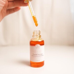 K&ouml;nnte beinhalten: Eine durchsichtige Glasflasche mit goldenem Deckel und einer Pipette, gef&uuml;llt mit orangefarbener Fl&uuml;ssigkeit. Das Flaschenetikett lautet "Glow Facial Serum".