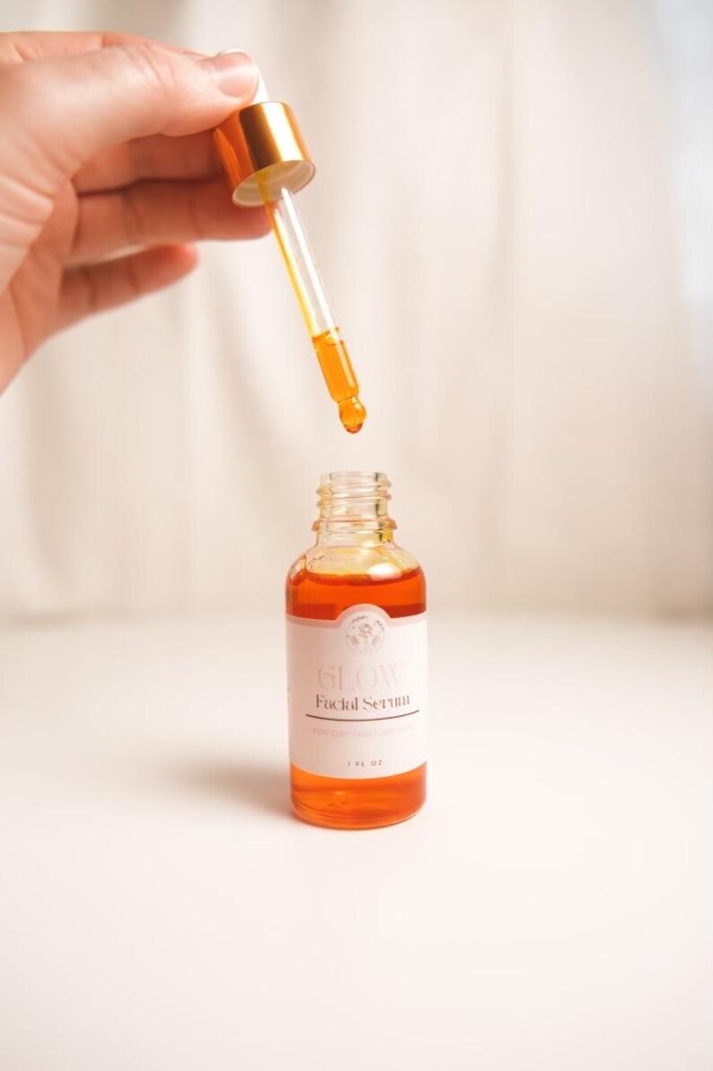 K&ouml;nnte beinhalten: Eine durchsichtige Glasflasche mit goldenem Deckel und einer Pipette, gef&uuml;llt mit orangefarbener Fl&uuml;ssigkeit. Das Flaschenetikett lautet "Glow Facial Serum".