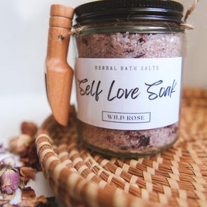 May include: A glass jar filled with pink bath salts with a wooden scoop attached to the side. The jar has a white label that reads "Herbal Bath Salts Self Love Soak Wild Rose".