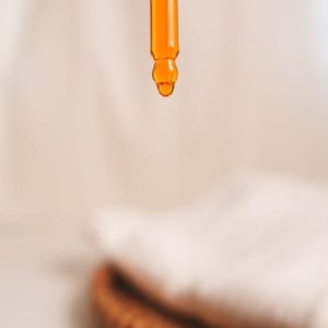 K&ouml;nnte beinhalten: Nahaufnahme eines Glas-Tropfers mit einer goldgelben Fl&uuml;ssigkeit, die von der Spitze tropft. Der Tropfer befindet sich vor einem wei&szlig;en Hintergrund.